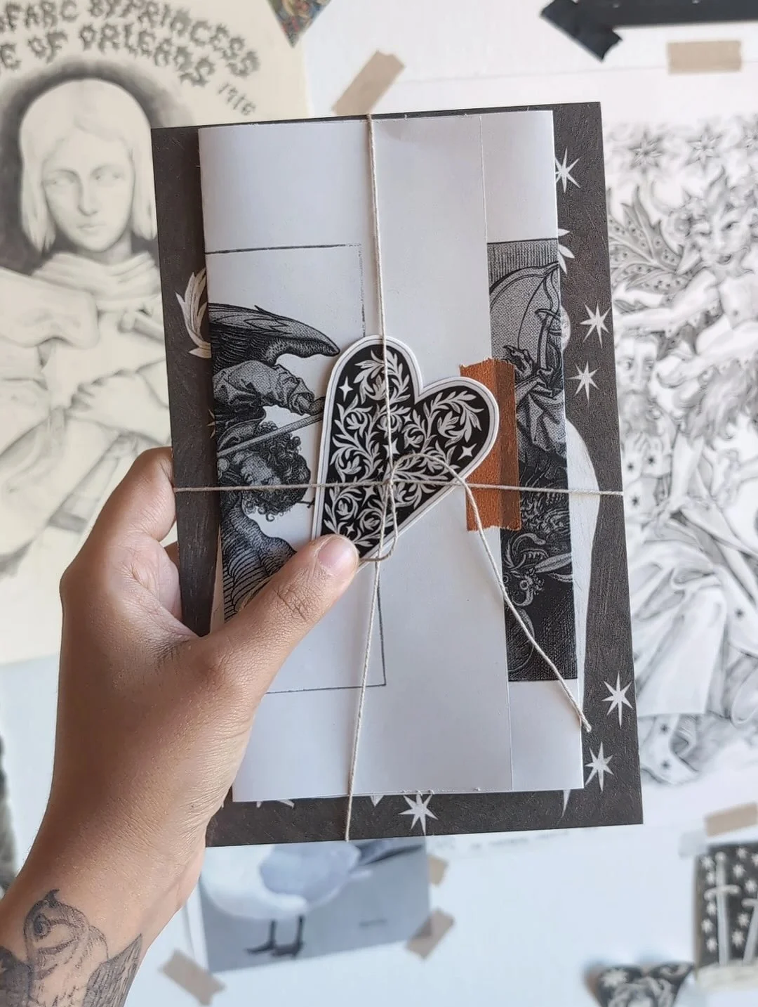 A hand holding a Peregrine Post bundle of black-and-white illustrated papers tied with string and an ornate heart sticker.