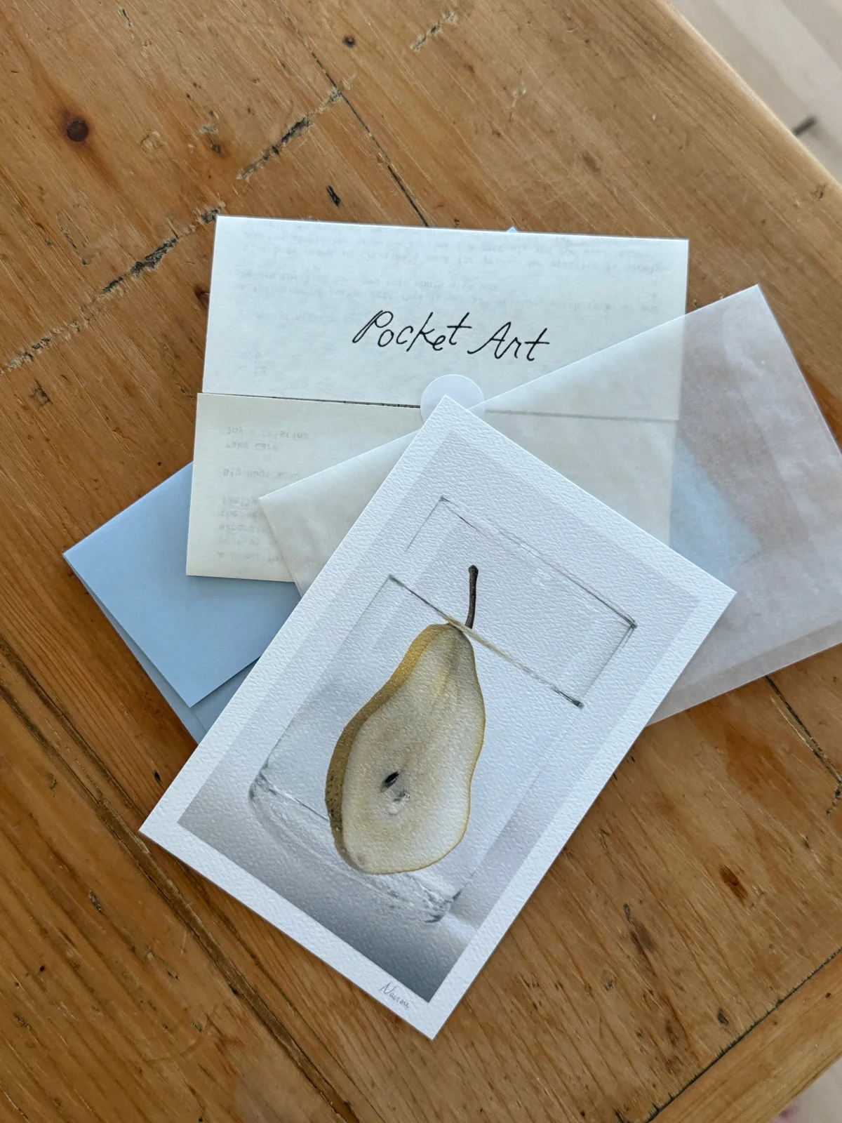 A pear print with a pale blue Pocket Art letter and envelope on a wooden table.